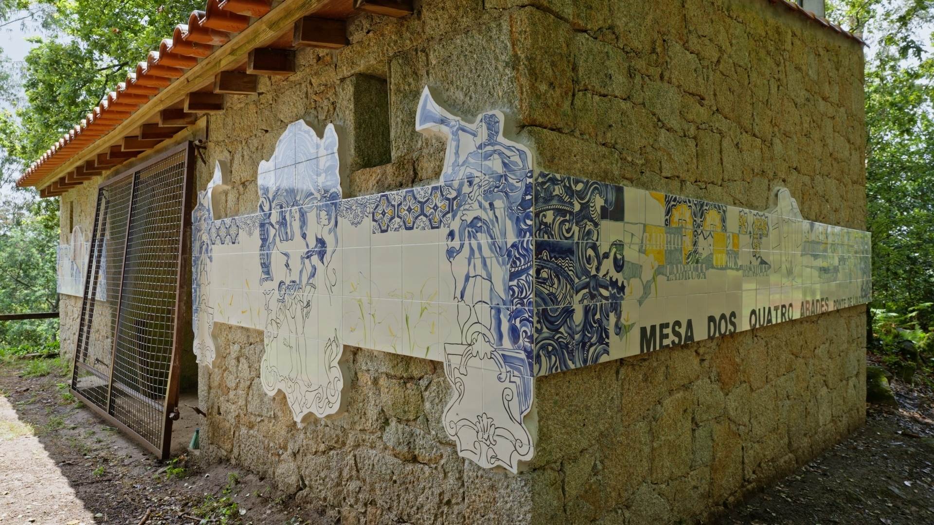 19.06.2022
The Mesa dos Quatro Abades (Table of Four Abbots) is a place for the collective memory of the people of Ponte de Lima, to which the roads of several parishes of this municipality converge. The artist was challenged to intervene in a pre-existing architectural structure, and chose to execute a tile panel. Given its characteristics, the set was divided into three parts.
The part facing the road deals with the issue of institutional cartography, represented by the table, the benches and a sketch of the compass rose. The names of all the parishes appear as if they were interventions in the urban space with mouldings. The whole top part departs from the idea of the traditional tile (15x15cm), despite being painted on a 20x20cm tile. This top is inspired by the carpet tiles that cover part of the facades and panelling of local churches. As it gets lower we move from traditional to contemporary, from excess to simplicity.
In the central part is the figure of Saint Sebastian, a figure that each of the parishes carried in procession to the Mesa dos Quatro Abades (Table of the Four Abbots). The figure of Saint Sebastian is flanked by two angels playing the trumpets, alluding to religious matters. The last part refers to more popular issues, such as pilgrimage and pastoralism.